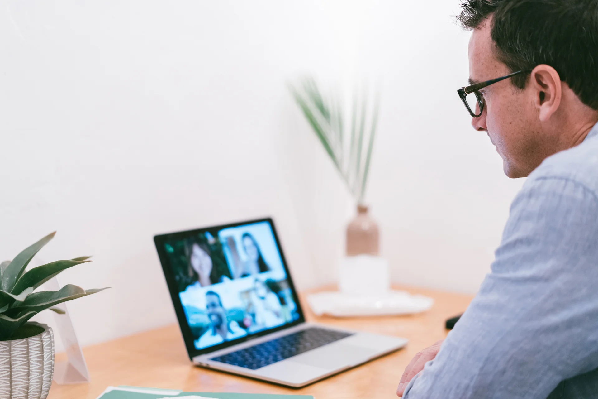 Professional Virtual Meeting with Focused Individual A man wearing glasses engaged in a video conference on a laptop at a neatly arranged wooden desk with a plant and decorative elements.